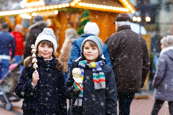 Petite fille et garçon mignon mangeant des fraises et des pommes couvertes de chocolat blanc sur brochette sur le marché de Noël allemand traditionnel. Enfants heureux, meilleurs amis, jumeaux et frères et sœurs le jour neigeux — Photo