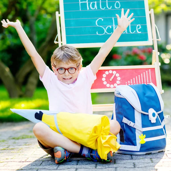Χαρούμενο αγοράκι με γυαλιά που κάθεται δίπλα στο γραφείο και σακίδιο ή σακίδιο. Μαθητής με παραδοσιακή γερμανική σχολική τσάντα που ονομάζεται Schultuete την πρώτη του μέρα στο σχολείο. Γεια σας σχολείο στη γερμανική γλώσσα — Φωτογραφία Αρχείου
