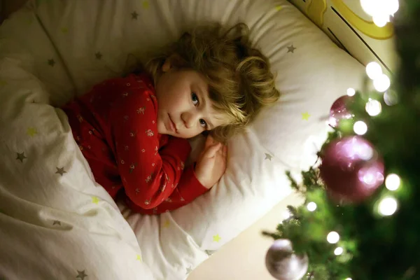 Petite fille mignonne tout-petit au lit sous l'arbre de Noël et rêvant du Père Noël à la maison, à l'intérieur. Fête chrétienne traditionnelle. Enfant heureux attendant des cadeaux pour Noël. Lumière douce et confortable — Photo