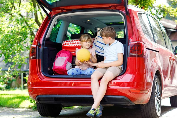 Duas crianças, menino da escola e menina pré-escolar sentados no porta-malas do carro antes de sair para férias de verão com os pais. Crianças felizes, irmãos, irmão e irmã com malas e brinquedos em viagem — Fotografia de Stock