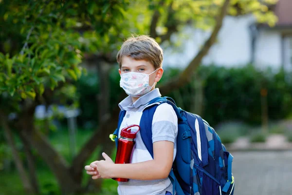 Rapazinho feliz, máscara médica, garrafa de água e mochila ou mochila. Um estudante a caminho da escola. Criança saudável ao ar livre. De volta à escola após o tempo de quarentena do bloqueio da doença pandêmica da corona — Fotografia de Stock