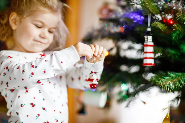 Adorabile bambina in pigiama che decora l'albero di Natale con un giocattolo in mani carine. Bambino in camicia da notte accanto all'albero di Natale. celebrazione della tradizionale vacanza invernale in famiglia — Foto Stock