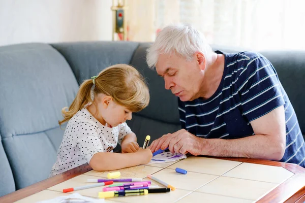 Schattig klein kleuter meisje en knappe senior grootvader schilderij met kleurrijke vilten pennen en potloden thuis. Kleinkind en man hebben plezier samen, creatieve familie. Indoor activiteit met kind. — Stockfoto