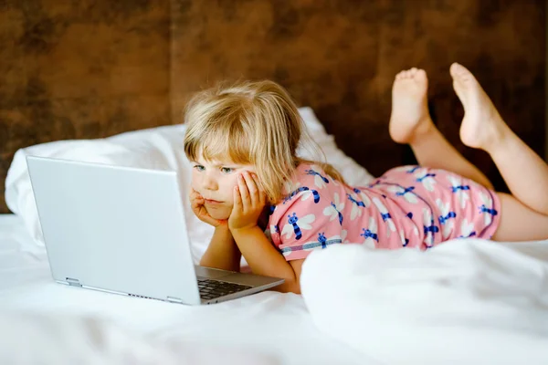 Förskola flicka tittar på videor på bärbar dator, anteckningsbok, i sängen på rena vita sängkläder. Aktivitet inomhus med barn. Frilans, distansundervisning eller arbete hemifrån med barn koncept. Lyckligt barn — Stockfoto