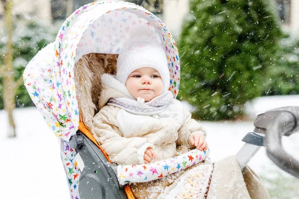 Schattig klein mooi baby meisje zittend in de kinderwagen of kinderwagen op koude besneeuwde winterdag. Gelukkig lachend kind in warme kleren, mode stijlvolle babyjas. Babys eerste sneeuw. Winterwandeling buiten. — Stockfoto