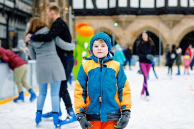 Renkli, kalın giysiler içinde, Noel pazarında ya da panayırda paten kayan mutlu küçük çocuk. Sağlıklı bir çocuk buz pateninde eğleniyor. Birçok insan bayramı kutluyor ve aktif kış tatili geçiriyor.