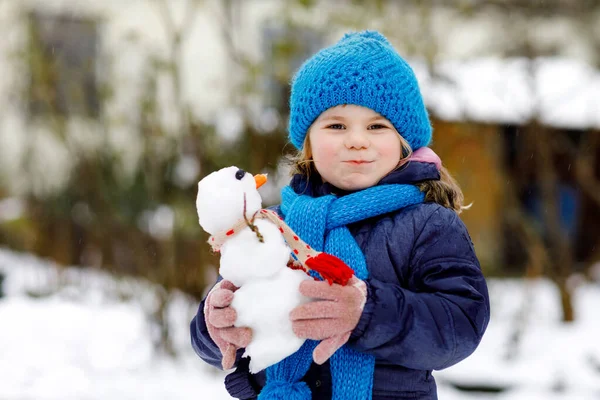 Χαριτωμένο κοριτσάκι που φτιάχνει μίνι χιονάνθρωπο και τρώει μύτη καρότου. Αξιολάτρευτο υγιές χαρούμενο παιδί που παίζει και διασκεδάζει με το χιόνι, σε εξωτερικούς χώρους την κρύα μέρα. Ενεργός ελεύθερος χρόνος με τα παιδιά το χειμώνα — Φωτογραφία Αρχείου
