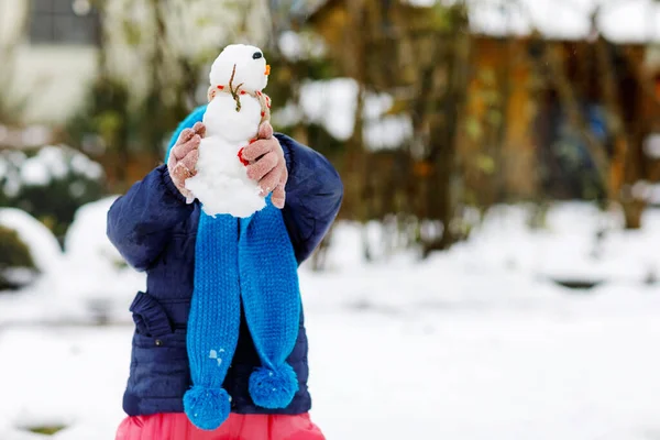 Cute little toddler girl making mini snowman and eating carrot nose. Adorable healthy happy child playing and having fun with snow, outdoors on cold day. Active leisure with children in winter — Stock Photo, Image