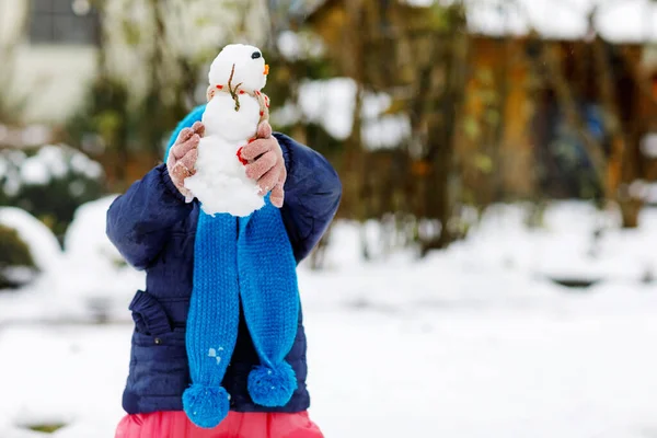 Linda niñita haciendo mini muñeco de nieve y comiendo la nariz de zanahoria. Adorable niño feliz saludable jugando y divirtiéndose con la nieve, al aire libre en el día frío. Ocio activo con niños en invierno —  Fotos de Stock