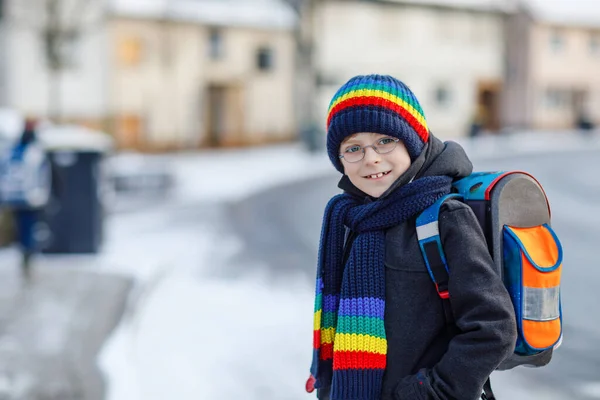 Chłopiec z podstawówki, idący do szkoły podczas opadów śniegu. Szczęśliwe zdrowe dziecko w okularach zabawy i zabawy ze śniegiem. Z plecakiem lub torbą w kolorowe zimowe ubrania. — Zdjęcie stockowe