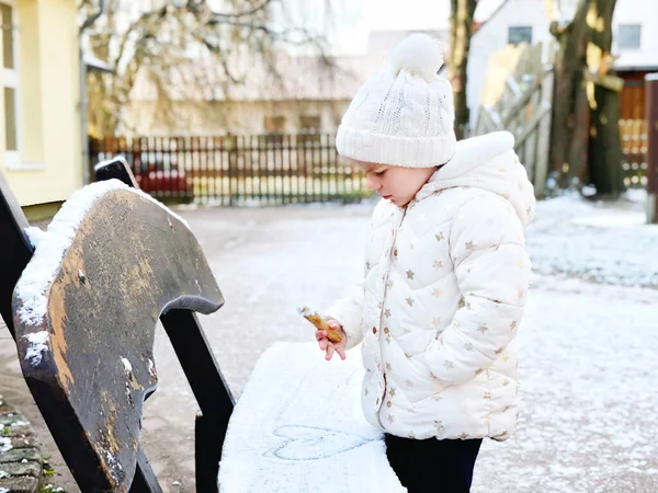 Little girl make walk outside and play with the first snow. Adorable happy child in winter clothes drawing heart on bench with snow. Kid during snowfall, outdoors. — Stock Photo, Image