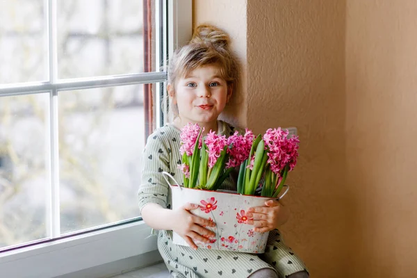 Piccola bambina seduta alla finestra con fiori di giacinto rosa in fiore. Bambino felice, in casa. Festa della mamma, San Valentino o compleanno e concetto di primavera. — Foto Stock