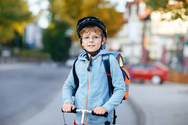 Active school kid boy in safety helmet riding with his scooter in the city with backpack on sunny day. Happy child in colorful clothes biking on way to school. — Stock Photo, Image