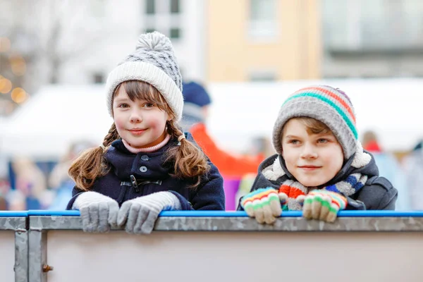 Dos niños pequeños y felices niña y niño en ropa caliente colorido patinaje en una pista de mercado de Navidad o feria. Niños sanos divirtiéndose en patinaje sobre hielo. Mucha gente que tiene ocio activo de invierno. — Foto de Stock