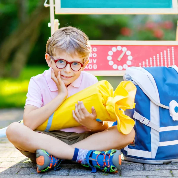 Gelukkige kleine jongen met een bril aan zijn bureau en rugzak of tas. Schoolkind met traditionele Duitse schooltas kegel genaamd Schultuete op zijn eerste dag naar school — Stockfoto