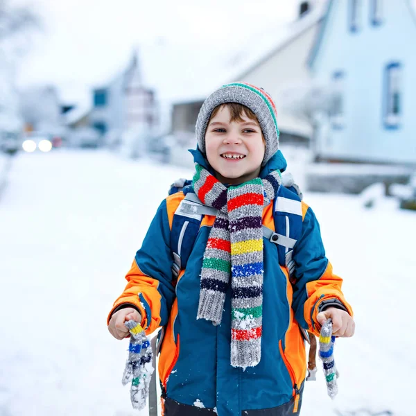 En liten skolpojke i grundskolan som går till skolan under snöfallet. Tidig morgon och snöiga gator i staden. Studerande med ryggsäck eller väska i färgglada vinterkläder. — Stockfoto