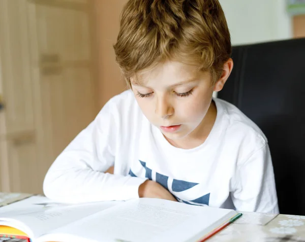 En blond skolpojke som läser en bok hemma. Barn intresserade av att läsa tidningen för barn. Fritid för barn, bygga färdigheter och utbildning koncept — Stockfoto