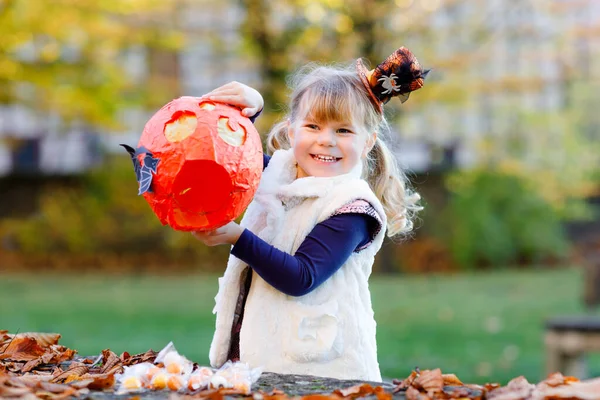 Kleines Mädchen, das sich als Hexe verkleidet oder an Halloween behandelt. Glückliches Kind im Freien, mit orangefarbener lustiger Mütze und Kürbisbeutel für süßen Spuk. Familienfestsaison im Oktober. Aktivitäten im Freien — Stockfoto