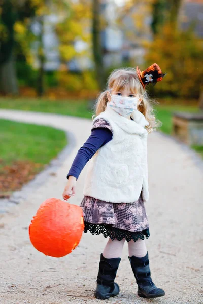 Маленькая девочка, одетая как ведьма с медицинской маской на лице трюк или лечить в Хэллоуин. Ребенок на улице, в смешной шляпе и сумке для сладких призраков. Время карантина вируса короны пандемии — стоковое фото