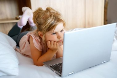 Anaokulundaki kız dizüstü bilgisayarda, defterde, yatakta temiz beyaz çarşaflarda videolar izliyor. Çocuklarla kapalı mekan aktivitesi. Serbest çalışma, uzaktan öğrenme ya da çocuk konseptiyle evden çalışma. Mutlu çocuk