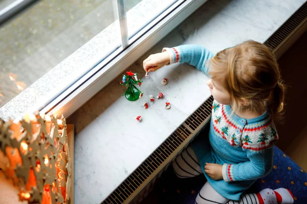 Menina pequena criança sentada à janela e decorando pequena árvore de Natal de vidro com minúsculos brinquedos xmas. A criança sã feliz celebra férias tradicionais de família. Adorável bebê. — Fotografia de Stock