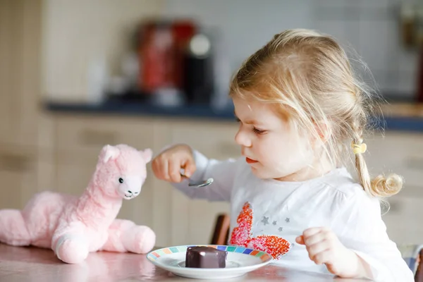 Menina engraçada e engraçada comendo sorvete de chocolate em casa. Criança bebê saudável feliz alimentando brinquedo de lama de pelúcia com sorvete doce. Criança adorável desfrutando de pudim de sobremesa. — Fotografia de Stock