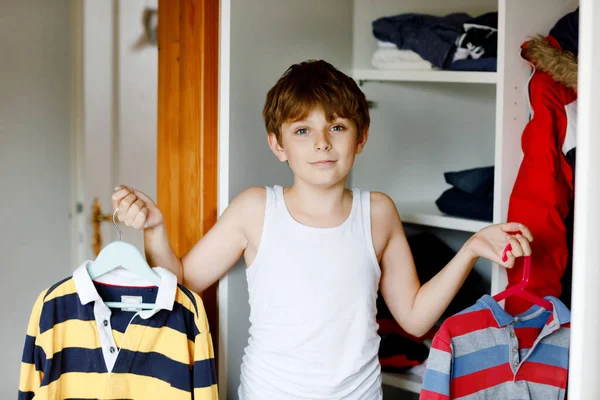 Παιδί του σχολείου στέκεται δίπλα στην ντουλάπα με ρούχα. Το παιδί αποφασίζει να φορέσει σχολική μπλούζα. Τα παιδιά ντύνονται το πρωί για το σχολείο.. — Φωτογραφία Αρχείου