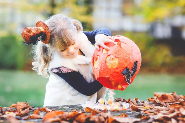 Niña pequeña vestida como un truco de bruja o trato en Halloween. Niño feliz al aire libre, con sombrero divertido naranja y bolsa de calabaza para dulce refugio. Temporada de festivales familiares en octubre. Actividades al aire libre —  Fotos de Stock