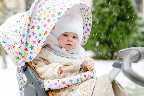 Schattig klein mooi baby meisje zittend in de kinderwagen of kinderwagen op koude besneeuwde winterdag. Gelukkig lachend kind in warme kleren, mode stijlvolle babyjas. Babys eerste sneeuw. Winterwandeling buiten. — Stockfoto