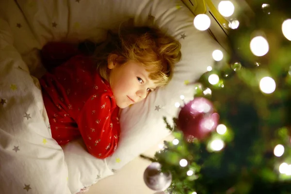 Petite fille mignonne tout-petit au lit sous l'arbre de Noël et rêvant du Père Noël à la maison, à l'intérieur. Fête chrétienne traditionnelle. Enfant heureux attendant des cadeaux pour Noël. Lumière douce et confortable — Photo