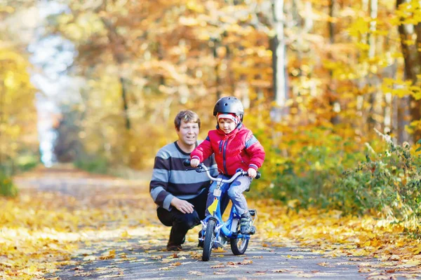 Маленький мальчик и его отец в осеннем парке на велосипеде. Папа учит своего сына кататься на велосипеде — стоковое фото