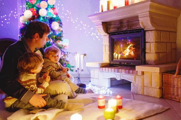 Vater und zwei kleine Jungen sitzen am Kamin, Kerzen und Kamin und schauen auf das Feuer. — Stockfoto