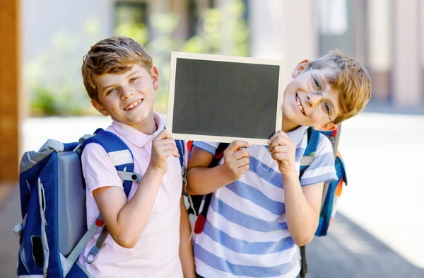 Två små pojkar med ryggsäck eller väska. Skolbarn på väg till skolan. Friska barn, bröder och bästa vänner utomhus på gatan lämnar hemmet. Skolor på kritskrivbordet. Lyckliga syskon. — Stockfoto