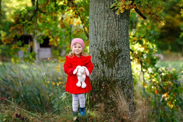 Beautiful toddler girl in red coat making a walk through autumn forest. Happy healthy baby holding soft toy rabbit. Sunny fall day with child. Active leisure and activity with kids in nature. — Stock Photo, Image