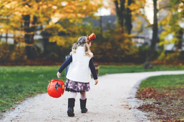 Little toddler girl dressed as a witch trick or treating on Halloween. Happy child outdoors, with orange funny hat and pumpkin bag for sweet haunt. Family festival season in october. Outdoor activity — Stock Photo, Image