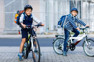 Kasklı iki okul çocuğu şehirde sırt çantalı bisikletle dolaşıyorlar. Renkli giysiler içinde okula bisikletle giden mutlu çocuklar. Çocuklar için güvenli okul yolu.