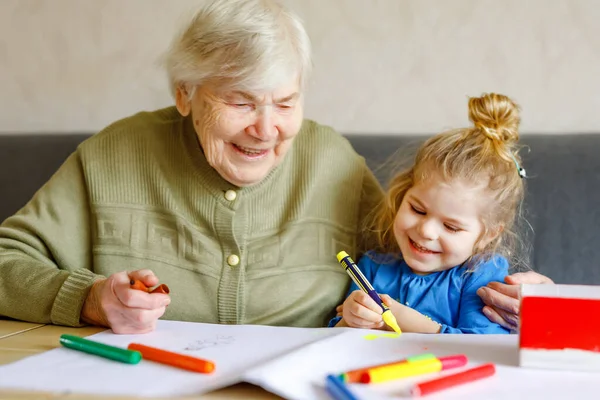 Όμορφο κοριτσάκι και γιαγιά ζωγραφίζει μαζί εικόνες με στυλό τσόχας στο σπίτι. Χαριτωμένο παιδί και ηλικιωμένη γυναίκα διασκεδάζουν μαζί. Ευτυχισμένη οικογένεια μέσα — Φωτογραφία Αρχείου