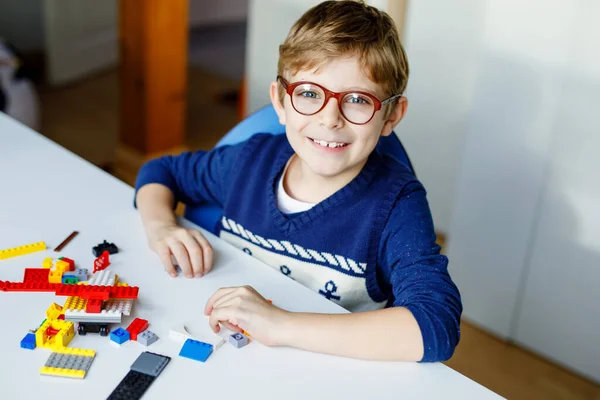 Malé blonďaté dítě s brýlemi si hraje se spoustou barevných plastových bloků. Rozkošný školní kluk baví s budováním a tvorbou robota. Kreativní volný čas moderní techniky a robotů. — Stock fotografie