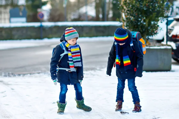 Twee kleine jongens van de basisklas die naar school lopen tijdens de sneeuwval. Gelukkige kinderen die plezier hebben en spelen met de eerste sneeuw. Broers en zussen en vrienden met rugzak in kleurrijke winterkleding. — Stockfoto