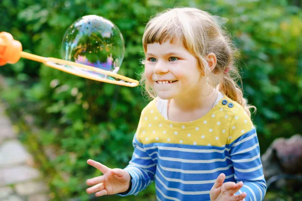 Happy little blonde preschool girl having fun with blowing soap bubble blower. Cute child playing on sunny summer day. Happy active funny healthy kid. Activity for children. — Stock Photo, Image