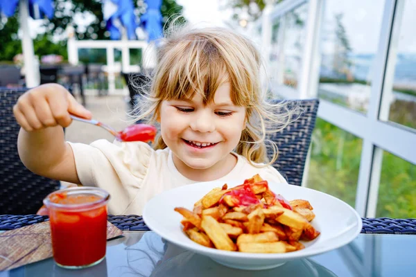 Porträt eines glücklich lächelnden Vorschulmädchens, das Pommes mit Tomatenketchup im Restaurant auf der Terrasse isst. Kleines Kind mit blonden Haaren genießt ungesundes Fast Food oder frisch zubereitetes Mittagessen. — Stockfoto