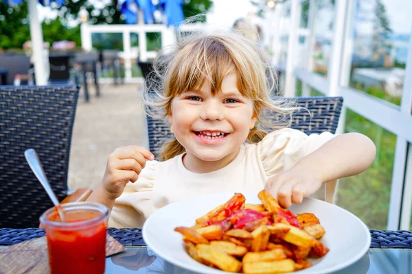 Портрет счастливой улыбающейся дошкольницы, которая ест картошку фри с томатным кетчупом в ресторане на террасе. Маленький ребенок с светлыми волосами наслаждается нездоровым фаст-фудом или свежим приготовленным обедом. — стоковое фото