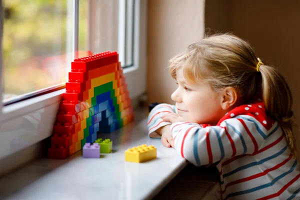 Милая маленькая девочка за окном создает радугу с красочными пластиковыми блоками во время пандемического карантина коронавируса. Дети делают и рисуют радуги по всему миру в знак. — стоковое фото