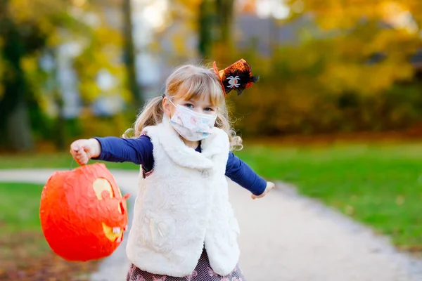 Маленькая девочка, одетая как ведьма с медицинской маской на лице трюк или лечить в Хэллоуин. Ребенок на улице, в смешной шляпе и сумке для сладких призраков. Время карантина вируса короны пандемии — стоковое фото