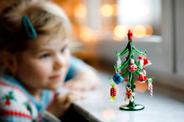 Menina pequena criança sentada à janela e decorando pequena árvore de Natal de vidro com minúsculos brinquedos xmas. A criança sã feliz celebra férias tradicionais de família. Adorável bebê. — Fotografia de Stock