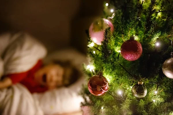 En söt liten flicka som sover under julgranen och drömmer om tomten hemma, inomhus. Traditionell kristen festival. Glada barn väntar på julklappar. Mysigt mjukt ljus — Stockfoto