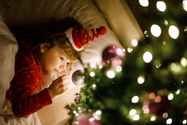 Petite fille mignonne tout-petit au lit sous l'arbre de Noël et rêvant du Père Noël à la maison, à l'intérieur. Fête chrétienne traditionnelle. Enfant heureux attendant des cadeaux pour Noël. Lumière douce et confortable — Photo