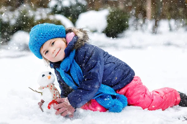 Küçük tatlı kız küçük kardan adam yapıyor ve havuç burnu yiyor. Soğuk bir günde dışarıda, karla oynayıp eğlenen sevimli, sağlıklı bir çocuk. Kışın çocuklarla aktif eğlence. — Stok fotoğraf