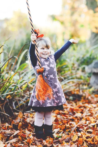 Linda niña pequeña vestida como una bruja celebra Halloween. Niño feliz al aire libre, con sombrero naranja divertido y escoba de bruja. Hermosa temporada de festivales familiares en octubre. Actividades al aire libre —  Fotos de Stock