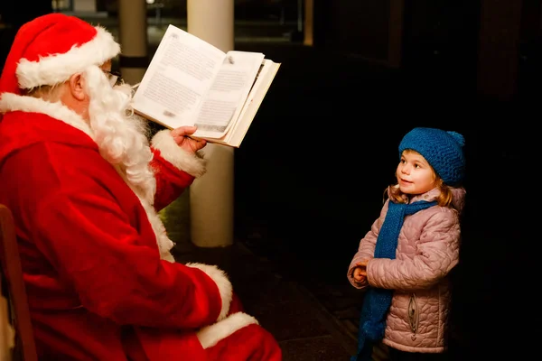 Little toddler girl talking to Santa Claus called Nikolaus or Weihnachtsmann in German. Happy smiling kid waiting for gifts. Santa with golden book. Xmas, childhood, holiday — Stock Photo, Image